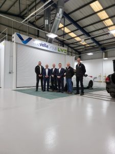 The Vella Group and LV= General Insurance colleagues stand at The Vella Group Bradford repair centre in front of a spraybooth that says 'The Vella Group working with LV='